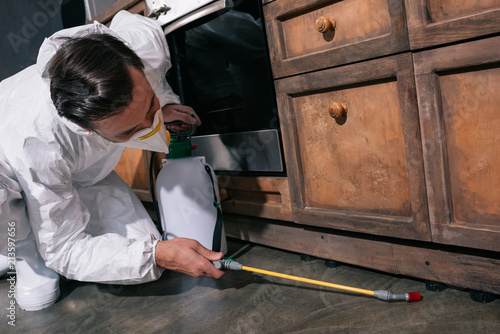 pest control worker in uniform spraying pesticides under cabinet in kitchen