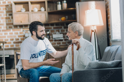 Nice talk. Good-looking inspired bearded volunteer smiling and talking with an old man while sitting in the kitchen