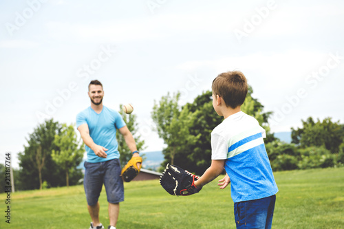 Handsome dad with his little cute sun are playing baseball on green grassy lawn