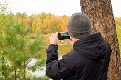 Young man in black jacket and gray knitted hat is taking a photo of autumn landscape on mobile phone in a forest near to the river, back view.