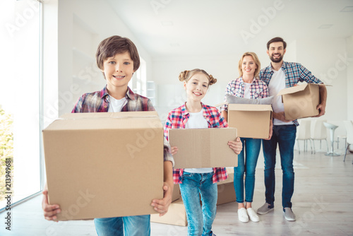 Young happy smiling family four persons wearing casual holding carton boxes with stuff things in light studio living room kitchen, moving to new flat