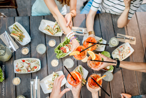 Friends clinking glasses with cocktail drinks, top view on the table full of snacks outdoors