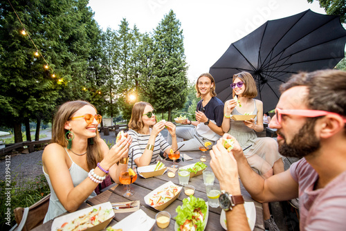 Young friends having fun sitting together with tasty snacks and drinks during the evening lights at the park cafe outdoors