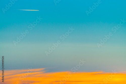 Sunrise landscape with airplane flying across blue sky and leave trail in clear sky above long orange cloudy line. Dawny cloudscape with copy space.