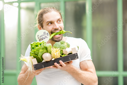 Portrait of a happy vegetarian man holding box full of fresh raw vegetables biting pepper outdoors on the green background