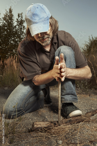Man in hat trying to make a fire with wood stick friction
