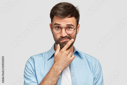 Image of hesitant unshaven European male with thick beard, holds chin, purses lips with clueless expressions, doubts what to eat delicious for supper, isolated over white background. What to choose?