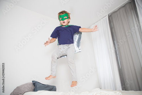 Cheerful little boy in Superhero's costume jumping on bed at home.