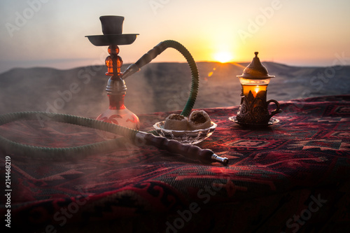 Hookah hot coals on shisha bowl making clouds of steam at desert outdoor. Oriental ornament on the carpet eastern tea ceremony. Stylish oriental shisha on sunset background.