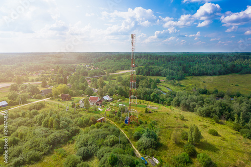 Telecommunication tower with radio antennas and satellite dishes is installed on the rural on the green field with grass, bushes and trees. Concept of harmless of electromagnetic and microwav