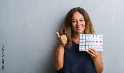 Middle age hispanic woman standing over grey grunge wall holding period calendar pointing and showing with thumb up to the side with happy face smiling