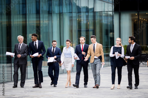 Several business people in elegant suits walking by modern building while having talk with each other