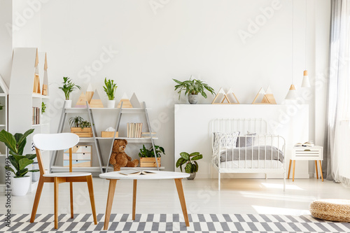 Wooden chair and table with open book placed on carpet in real photo of bright Scandinavian style bedroom interior for teenager with metal bed, fresh plants and triangle racks