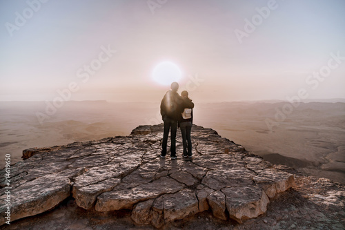 Alone young couple of man and women with backpack in israel negev desert admires the view of sunrise. Young pair stands on the edge of the cliff of makhtesh ramon park.