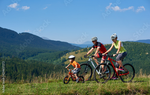 Sporty active family cyclists, mother, father and kid cycling on bikes on grassy hill. Carpathian mountains, blue summer sky on background. Healthy lifestyle, traveling and happy relations concept.