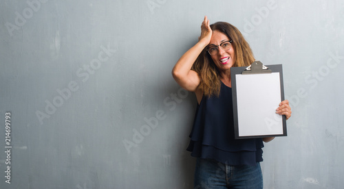 Middle age hispanic woman standing over grey grunge wall holding clipboard stressed with hand on head, shocked with shame and surprise face, angry and frustrated. Fear and upset for mistake.