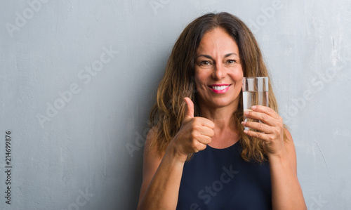 Middle age hispanic woman drinking glass of water happy with big smile doing ok sign, thumb up with fingers, excellent sign