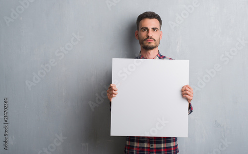 Young adult man over brick wall holding banner with a confident expression on smart face thinking serious