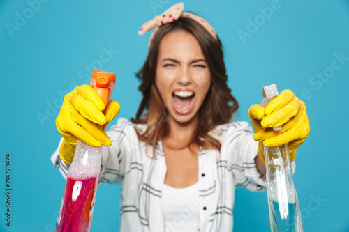 Photo closeup of displeased upset woman 20s in yellow rubber gloves for hands protection screaming while holding two detergent sprayers during cleaning, isolated over blue background