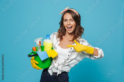 Photo of pleased young housemaid 20s wearing yellow rubber gloves for hands protection holding bucket with cleaning supplies, isolated over blue background