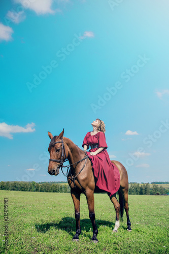 Beautiful girl in a long red dress riding a brown horse against a blue sky. The girl looked up and looked at the sky.
