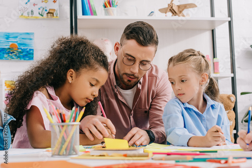 teacher in eyeglasses helping multicultural preschoolers with drawing at table in classroom