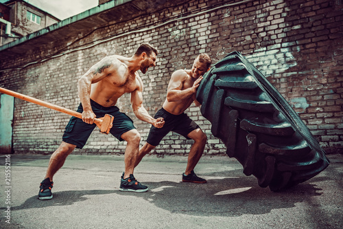 Two muscular athletes training. Muscular fitness shirtless man moving large tire other motivate him and hold big hummer in street gym. Concept lifting, workout training.