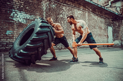 Two muscular athletes training. Muscular fitness shirtless man moving large tire other motivate him and hold big hummer in street gym. Concept lifting, workout training.