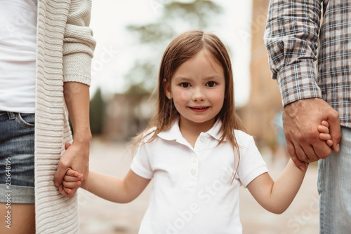 Waist up portrait of small smiling girl standing and holding mother and father hands. She is happy to spend time with dearest together