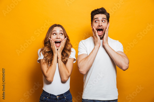 Photo of amazed couple man and woman in basic clothing screaming in surprise or delight and touching cheeks, isolated over yellow background
