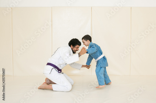 Father and little kid son are engaged in wrestling jiu-jitsu in the gym in a kimono. Trainer teaches child the methods and positions of single combat, karate or aikido.