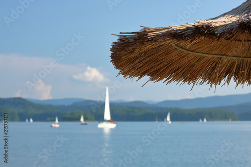 Plażowy parasol z trzciny, górna część, w tle błękitne niebo, jezioro z białymi żaglówkami na wodzie, rozmyte, wzgórza na drugim brzegu porośnięte gęstą roślinnością
