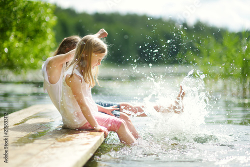Two cute little girls sitting on a wooden platform by the river or lake dipping their feet in the water on warm summer day