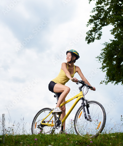 Attractive happy female cyclist cycling on yellow mountain bike, wearing helmet, enjoying summer day in the mountains. Outdoor sport activity, lifestyle concept