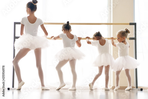 In a dance school, a group of girls and girls train together at the bar. all the little girls are dressed in white with gathered hair. Concept of: elegance, education, dance, learning