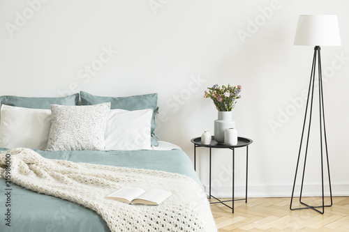 Real photo of bright bedroom interior with fresh flowers on bedside table, metal lamp and open book placed on bed with pastel bedding and knit blanket