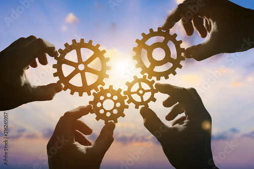 Four hands of businessmen collect gear from the gears of the details of puzzles. against the backdrop of dramatic sunlight. The concept of a business idea. Teamwork, strategy, cooperation, creativity