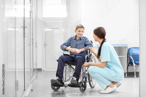 Young female doctor taking care of little boy in wheelchair indoors