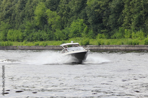 Travel, luxury water recreation on yacht - a white 19 ft motor boat fast gliding along the concrete shore of the navigable canal of Moscow in summer amid the trees on shore, day side view