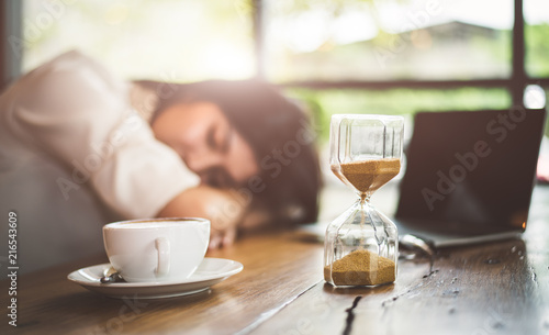 Close up of sandglass with business woman tired from working with laptop computer background. Relax Sleeping and taking nap concept. Business people and overtime workload theme. Office and home theme