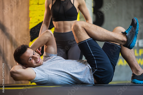 Cheerful fit guy is exercising obliques on mat in fitness studio. Ripped woman is kneeling behind him. Workout for health and beauty concept
