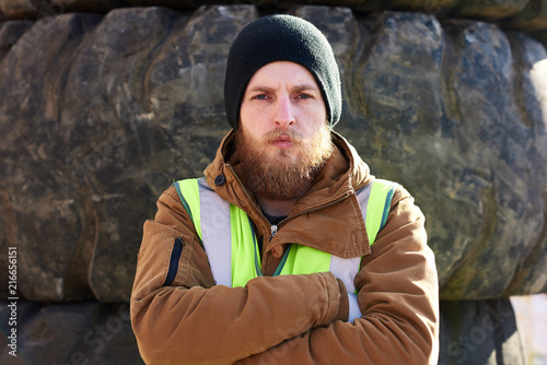 Portrait of tough bearded frontier worker standing with arms crossed and looking at camera against huge truck tires