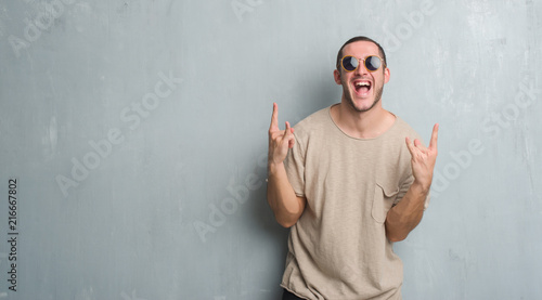 Young caucasian man over grey grunge wall wearing sunglasses shouting with crazy expression doing rock symbol with hands up. Music star. Heavy concept.