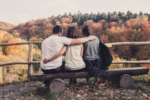 Three people hugged each other sitting on wooden benches and watch the beautiful autumn landscape, concept of polygamy