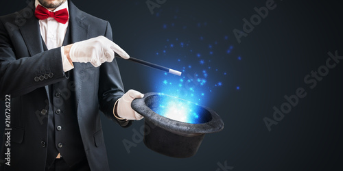 Magician or illusionist is showing magic trick with wand and hat on dark background.