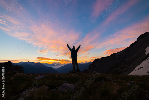 Man standing on mountain top outstretching arms, sunrise light colorful sky scenis landscape, conquering success leader concept.