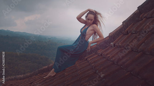 Aerial drone view of beautiful woman in long blue dress sitting on tiled red roof of the house against amazing mountain view and cloudy sky background