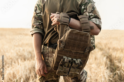 Soldier man standing against a field. Soldier in military outfit with bulletproof vest. Photo of a soldier in military outfit holding a gun and bulletproof vest on orange desert background.