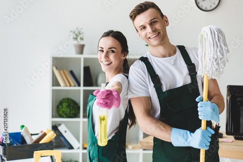 happy young cleaners with mop and spray bottle smiling at camera while cleaning office