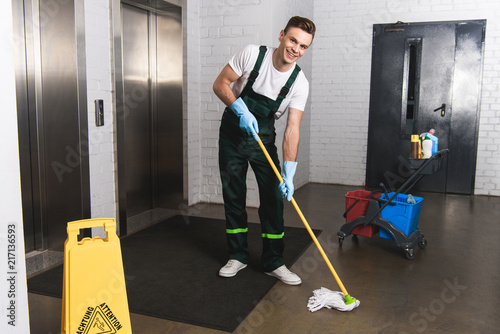 Handsome young janitor mopping floor and smiling at camera